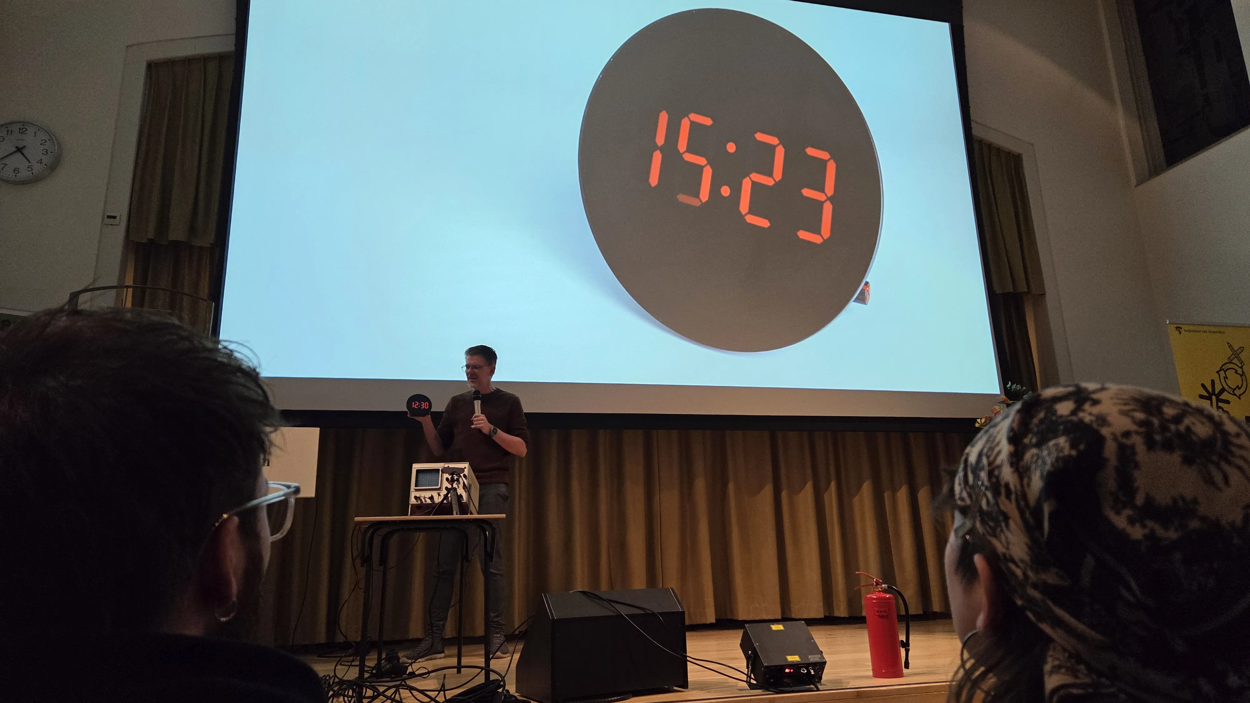 Man presenting a digital clock reading 12:30, with a larger projected image showing 15:23. Audience in foreground, screen and equipment visible.