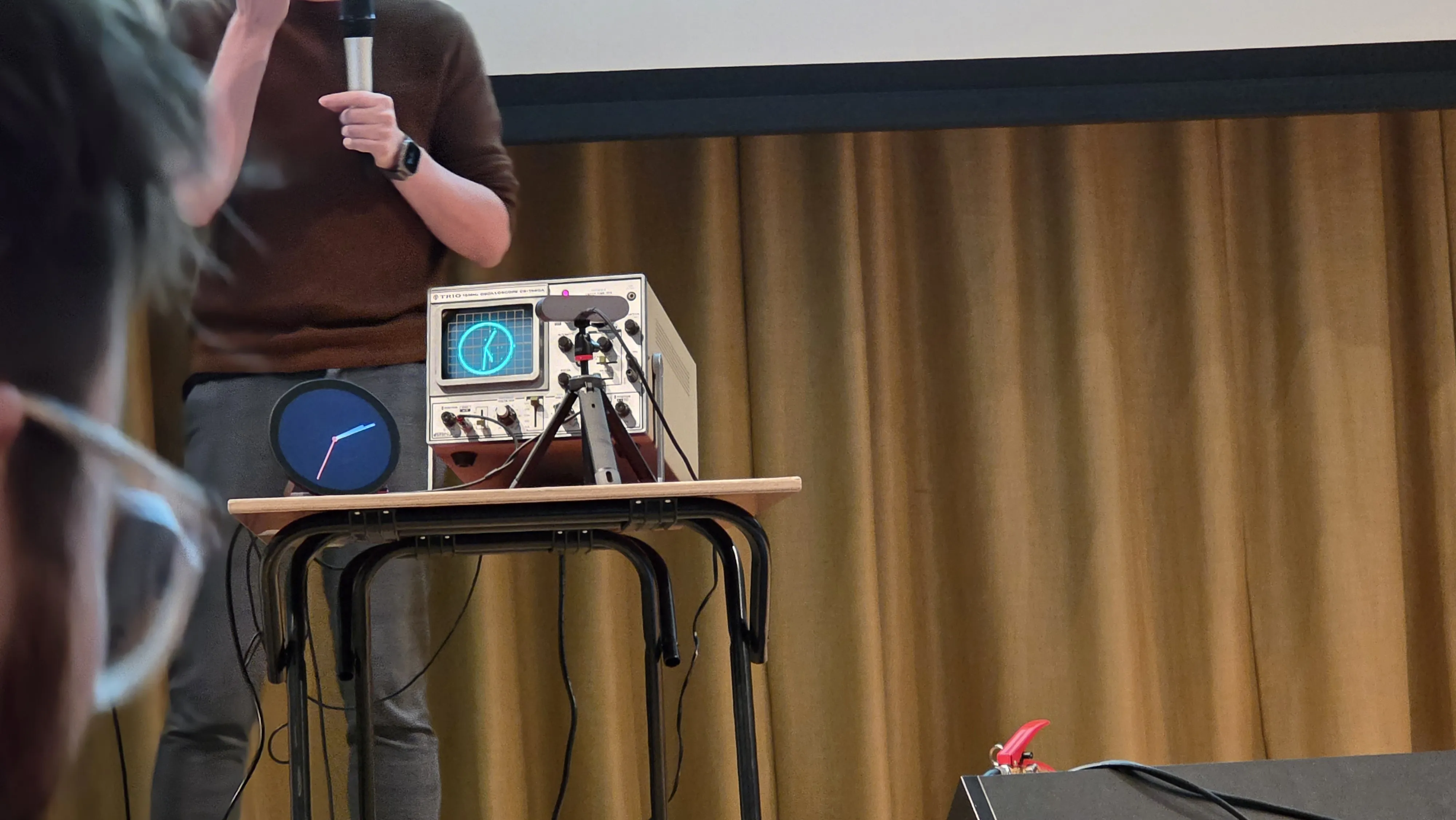 A person presenting with a microphone beside a tabletop oscilloscope and a clock, set against a gold curtain background.